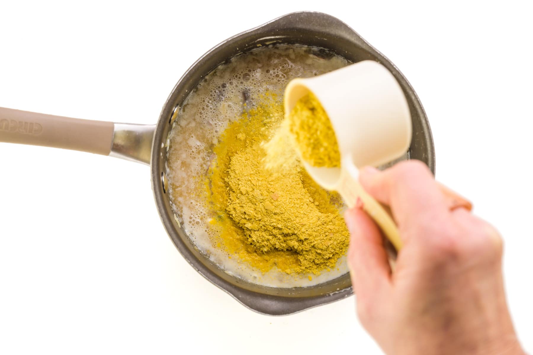 Nutritional yeast flakes are being poured into a saucepan with a thickened mushroom mixture.