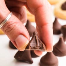 A hand holds a vegan chocolate kiss with a bite taken out. There are more of the candies and cookies in the background.