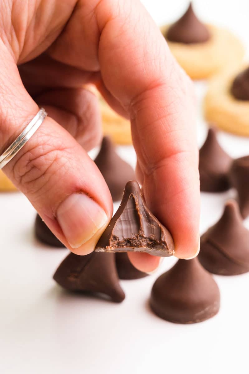 A hand holds a vegan chocolate kiss and takes a bite out.  There are more candies and cookies in the background.