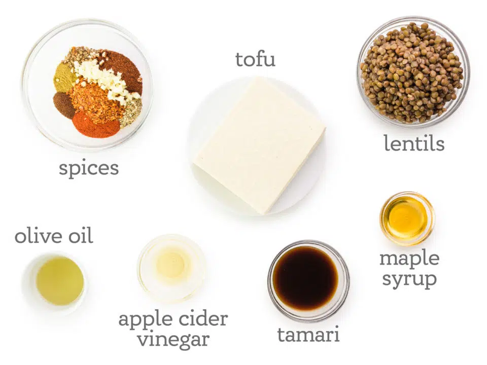 Ingredients are laid out on a table. The labels next to them read, lentils, maple syrup, tamari, apple cider vinegar, olive oil, spices, and tofu.