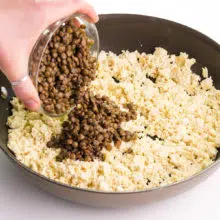 A hand holds a bowl of cooked lentls, pouring them into a skillet with tofu pieces.