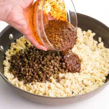 A hand is pouring a bowl of sauce into a skillet with tofu and lentils.
