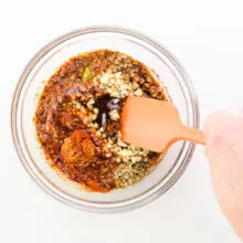 A hand holds a spatula stirring together sauce ingredients in a bowl.
