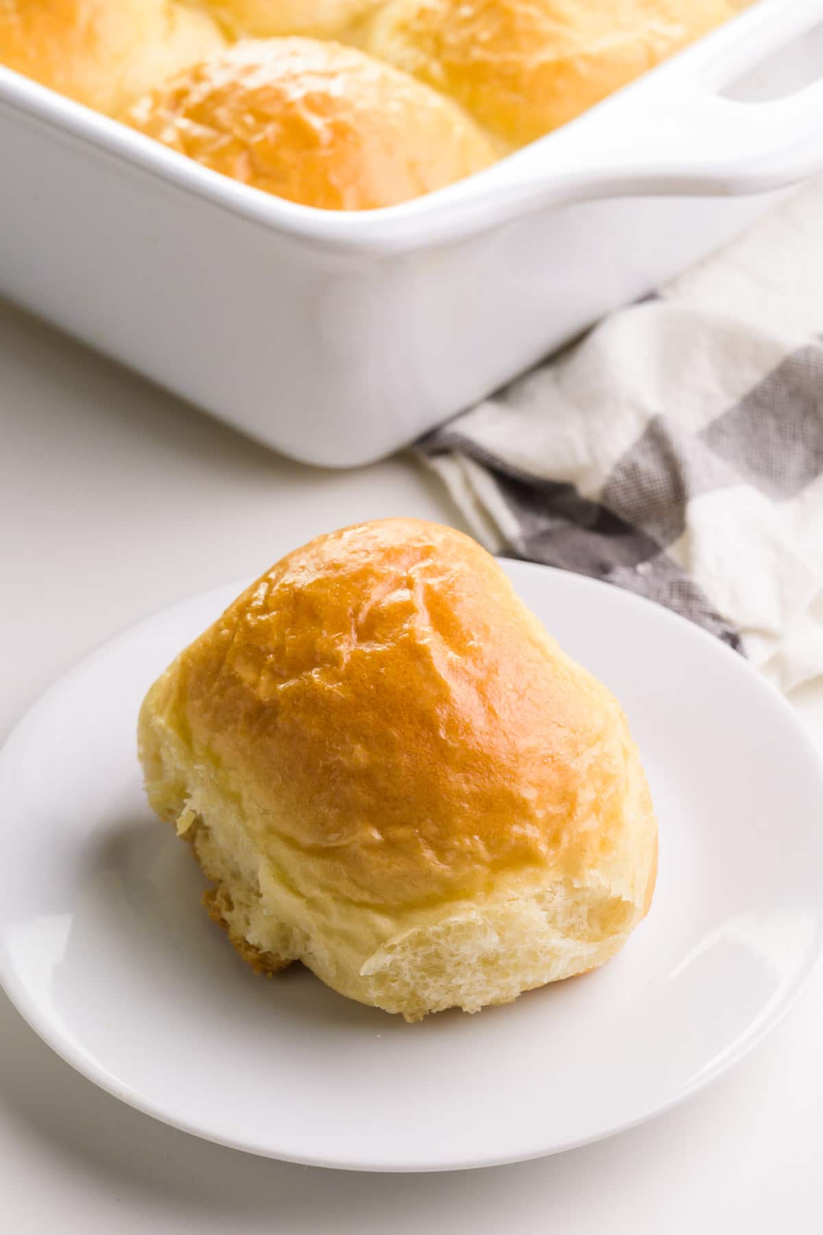 A vegan yeast roll sits on a plate in front of a pan with more rolls.