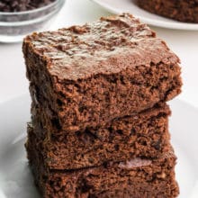 A stack of three gluten-free brownies sit on a plate in front of a bowl of chocolate chips ad more brownies in the background.