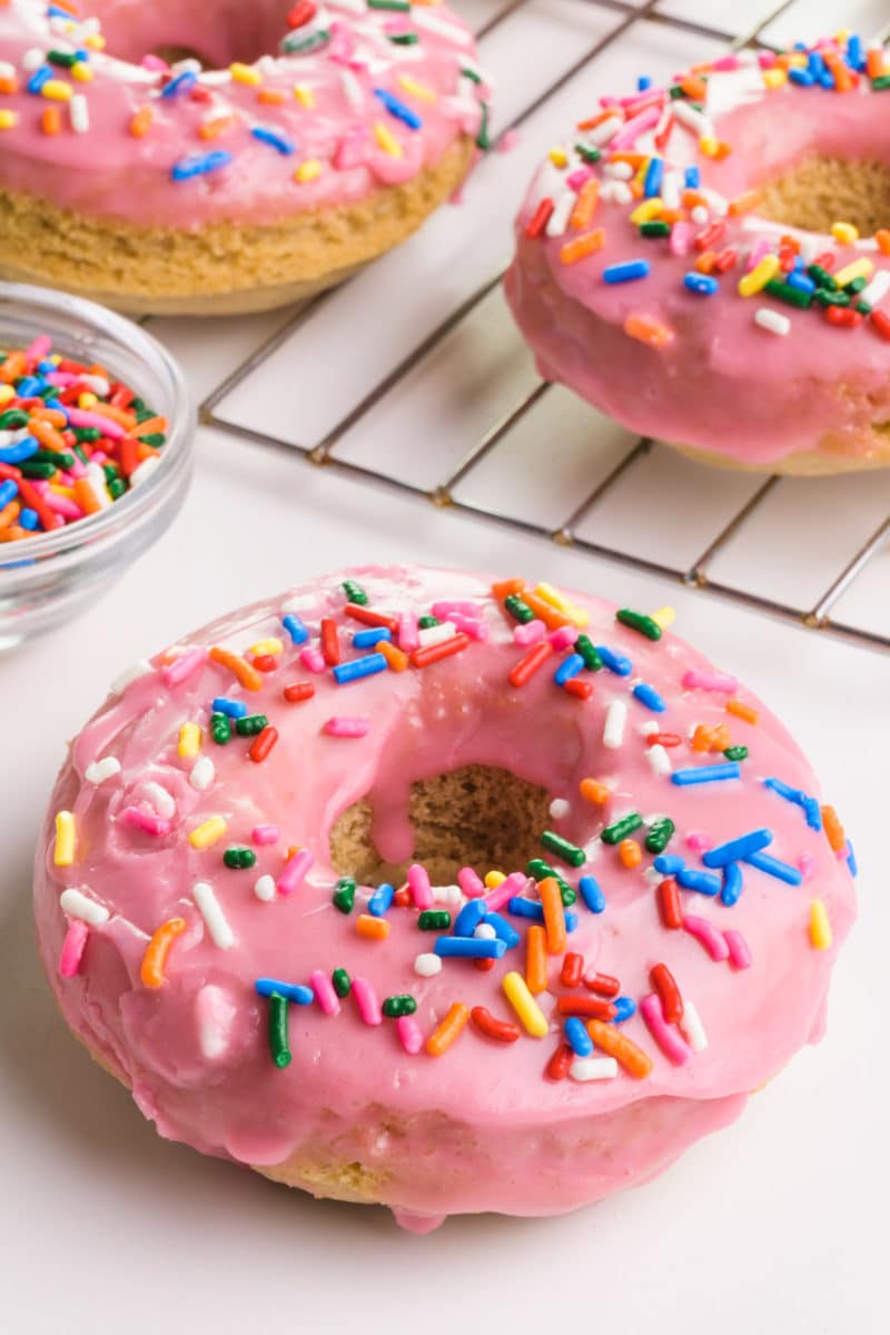 A gluten-free donut sits on a white table and a bowl of sprinkles in front of more donuts in the background.