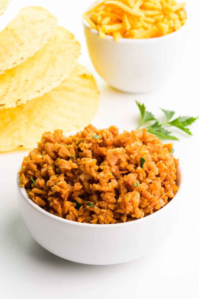 A bowl of vegan ground beef crumbles sits in front of taco shells, shredded cheese, and fresh herbs.