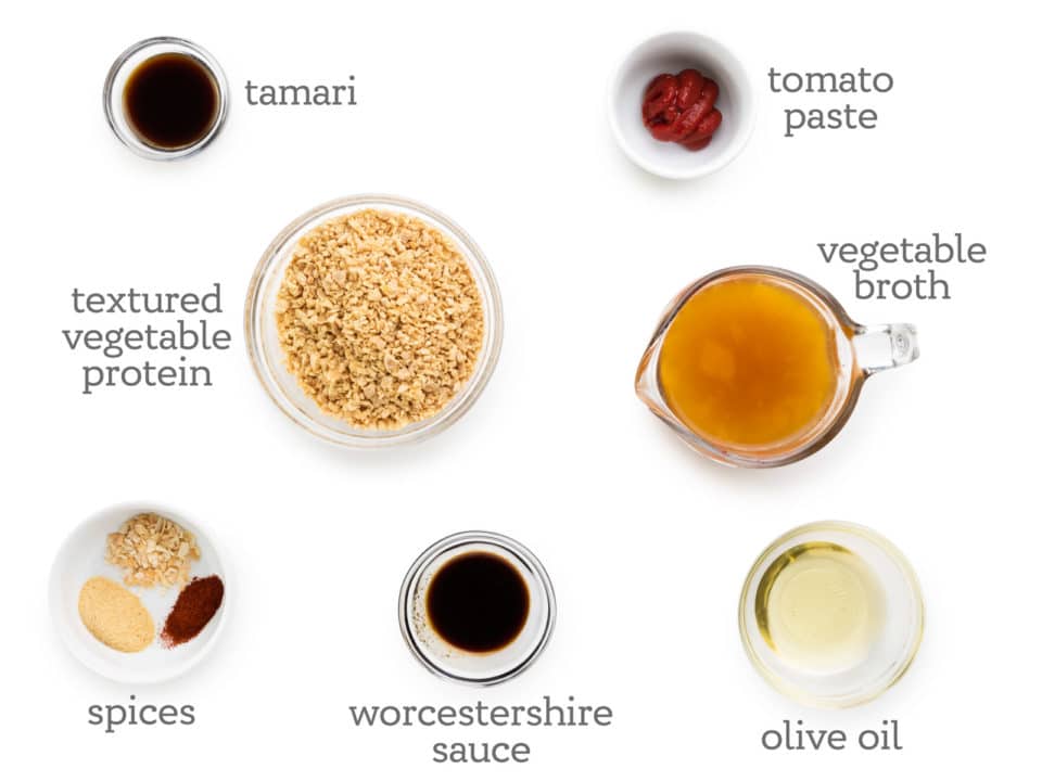 Ingredients are laid out on a white table. The labels next to them read, tomato paste, vegetable broth, olive oil, Worcestershire sauce, spices, textured vegetable protein, and tamari.
