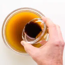 A hand holds a bowl with sauce, pouring it into another bowl with vegetable broth.