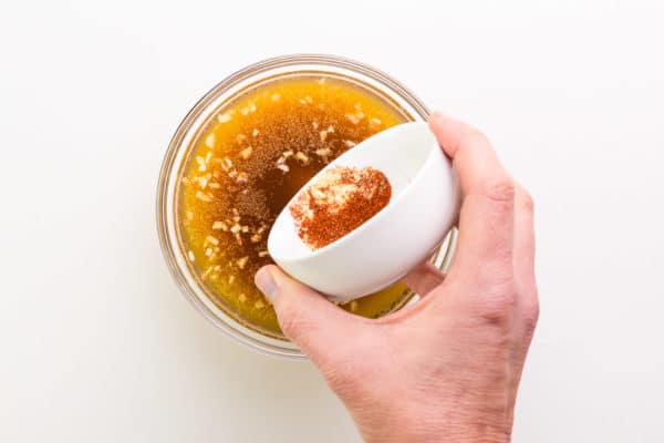 A hand holds a bowl of spices adding them to another bowl with a broth mixture.