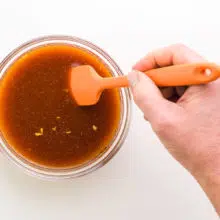 A hand holds a rubber spatula, stirring sauce in a bowl.