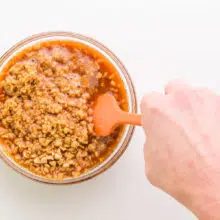 A hand holds a rubber spatula, stirring a bowl with sauce and textured vegetable protein.