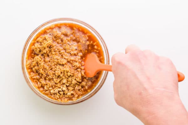 A hand holds a rubber spatula, stirring a bowl with sauce and textured vegetable protein.