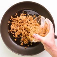 A hand holds a bowl of veggie crumbles, pouring it into a pan with olive oil.
