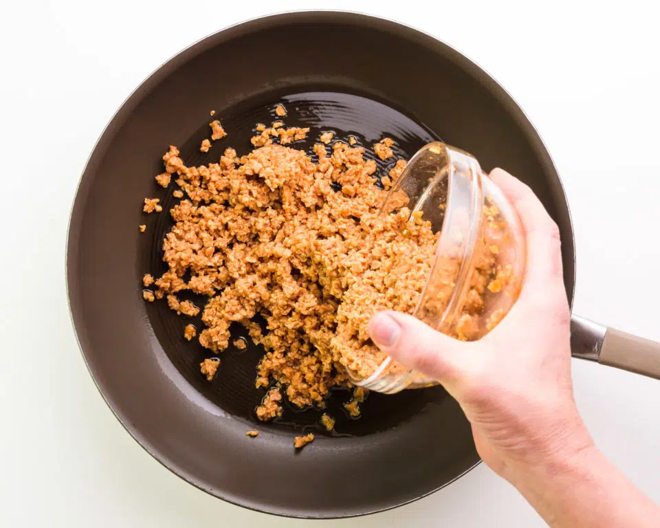 A hand holds a bowl of veggie crumbles, pouring it into a pan with olive oil.