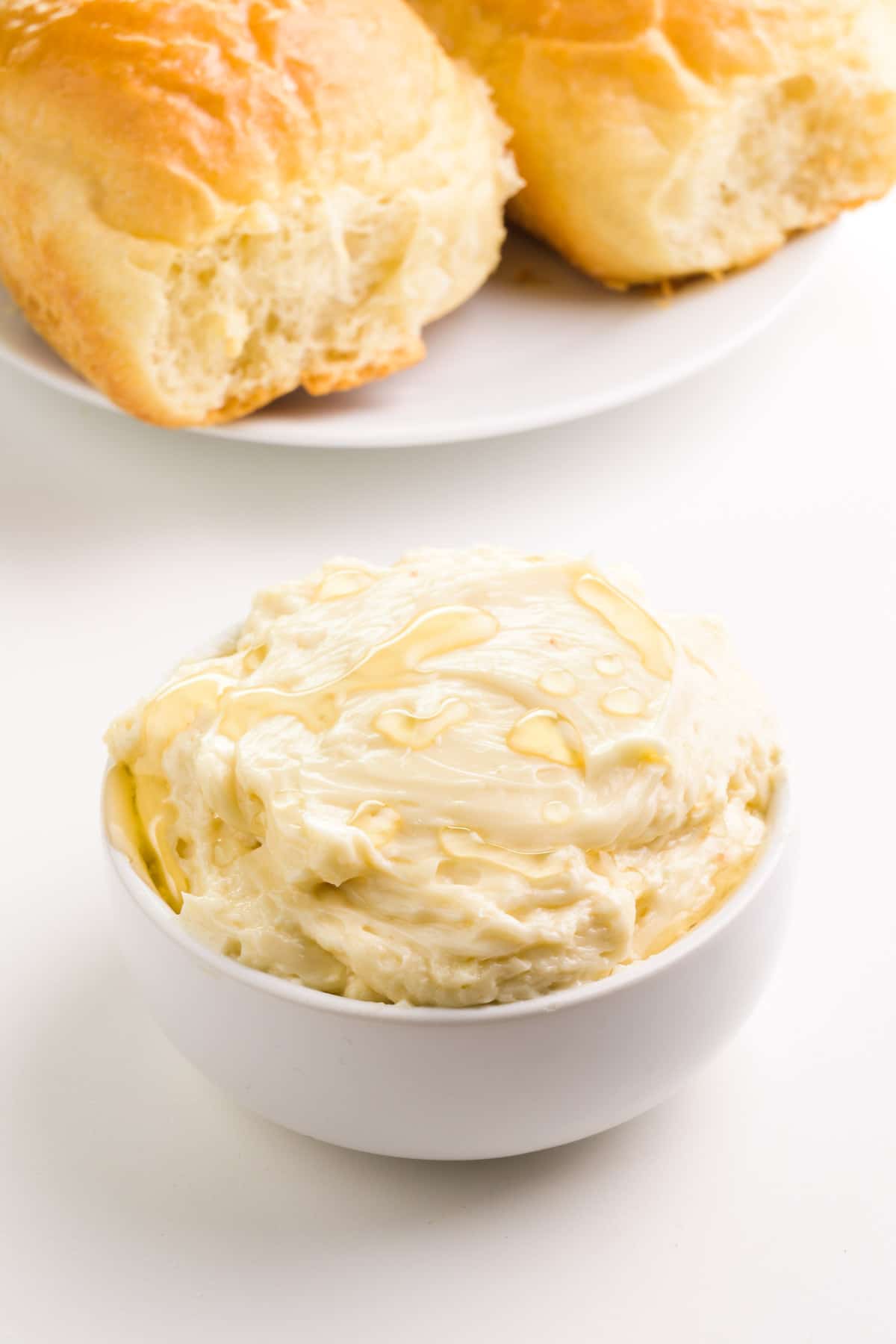 A bowl of vegan honey butter sits in front of a plate with dinner rolls.