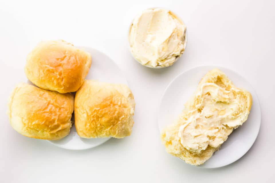 Looking down a dinner roll with vegan honey butter spread over the top. There's another bowl with more of the butter and a plate with three dinner rolls on it.