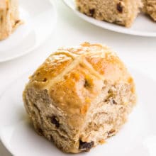 A vegan hot cross bun sits on a plate. There are more buns on plates in the background.