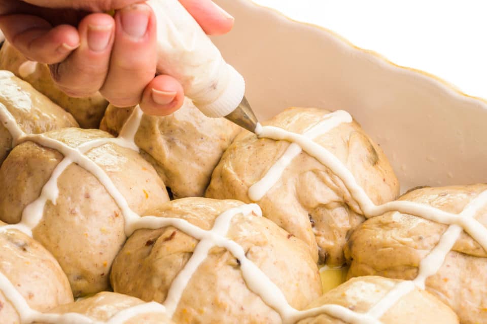 A hand holds a piping bag, piping crosses onto buns in a baking dish.