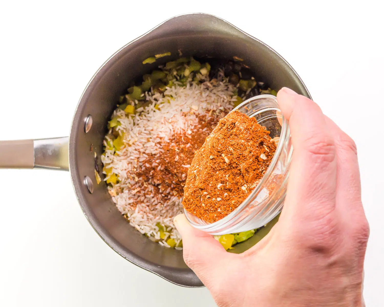 A hand holds a bowl of cajun seasoning, pouring it into a saucepan.