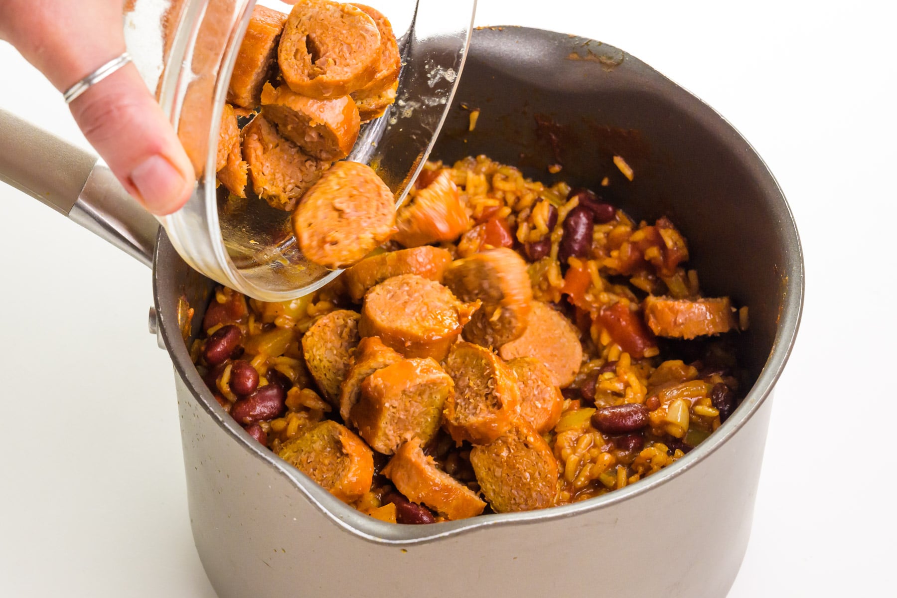 Sliced vegan sausage is being poured into a saucepan with tomato-based ingredients.