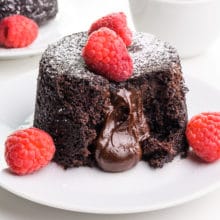 A vegan lava cake sits on a plate with chocolate coming out. There are fresh raspberries on top and beside it. Another plate is barely visible in the background.