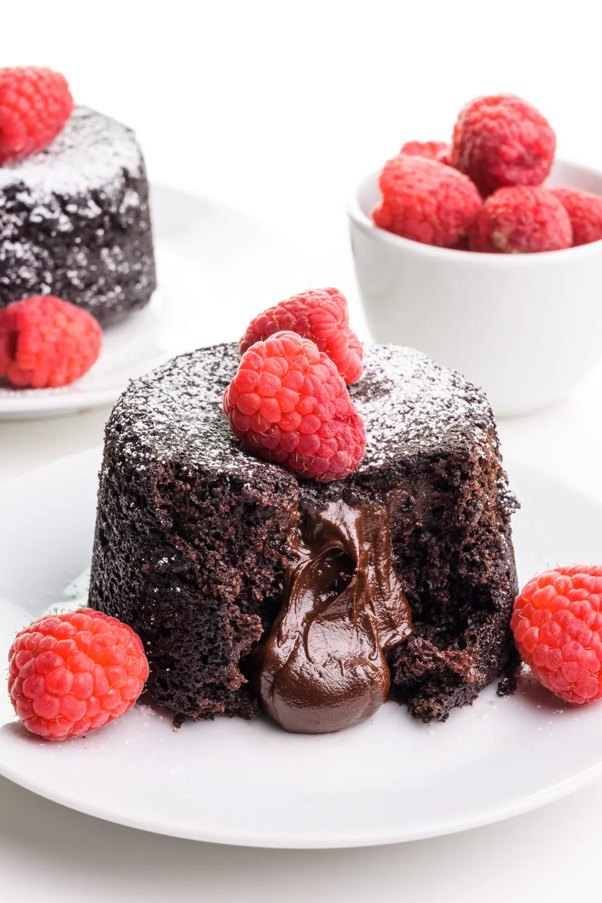 A vegan lava cake sits on a plate with molten chocolate coming out. There are fresh raspberries on and around it. There's another one on a plate in the background and a bowl of raspberries.
