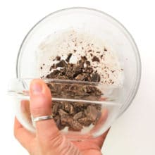 A hand holds a bowl of chopped chocolate, pouring it into a bowl with heated vegan creamer.