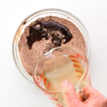 A hand holds a glass measuring cup full of liquid ingredients and is pouring it into a bowl with a flour and cocoa powder mixture.