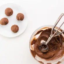Chocolate ganache balls sit on a white plate next to a bowl of chocolate with a cookie dispenser in it