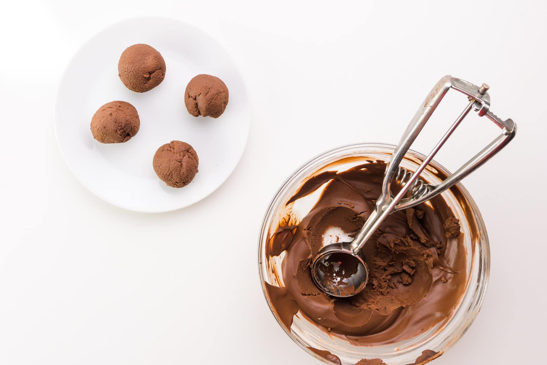 Chocolate ganache balls sit on a white plate next to a bowl of chocolate with a cookie dispenser in it