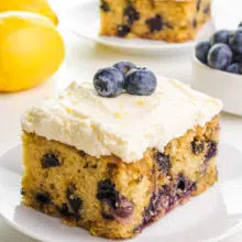 A lemon blueberry cake has frosting on top and fresh blueberries. There is a lemon in the background, another slice of cake, and a bowl of fresh blueberries.