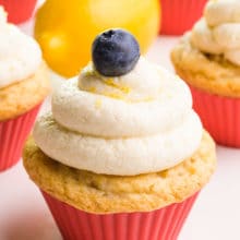 A cupcake has vegan lemon frosting on top with a blueberry and lemon zest. There are more cupcakes and a lemon in the background.