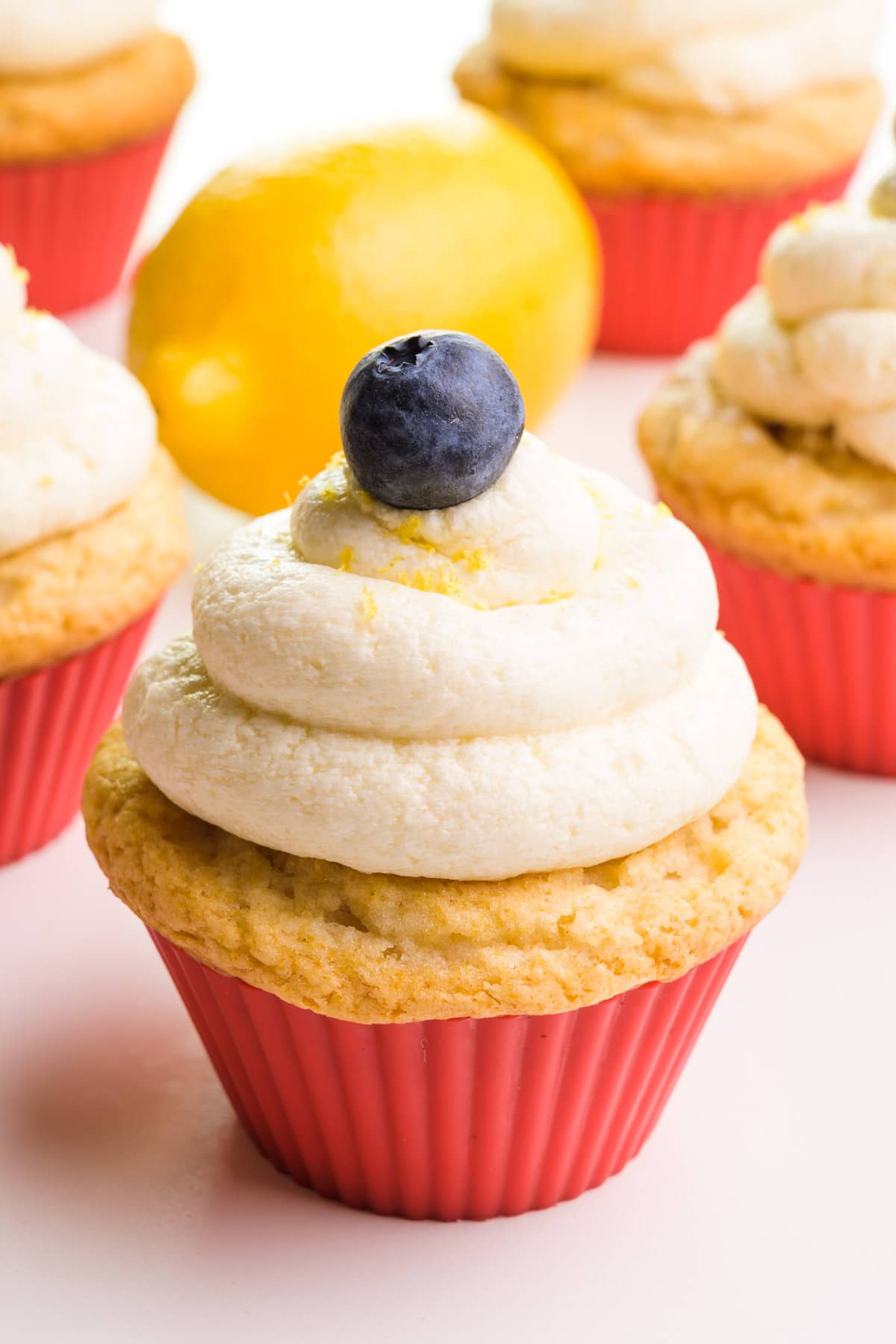 A cupcake topped with a blueberry and vegan lemon frosting with lemon zest.  In the background are more cupcakes and a lemon.