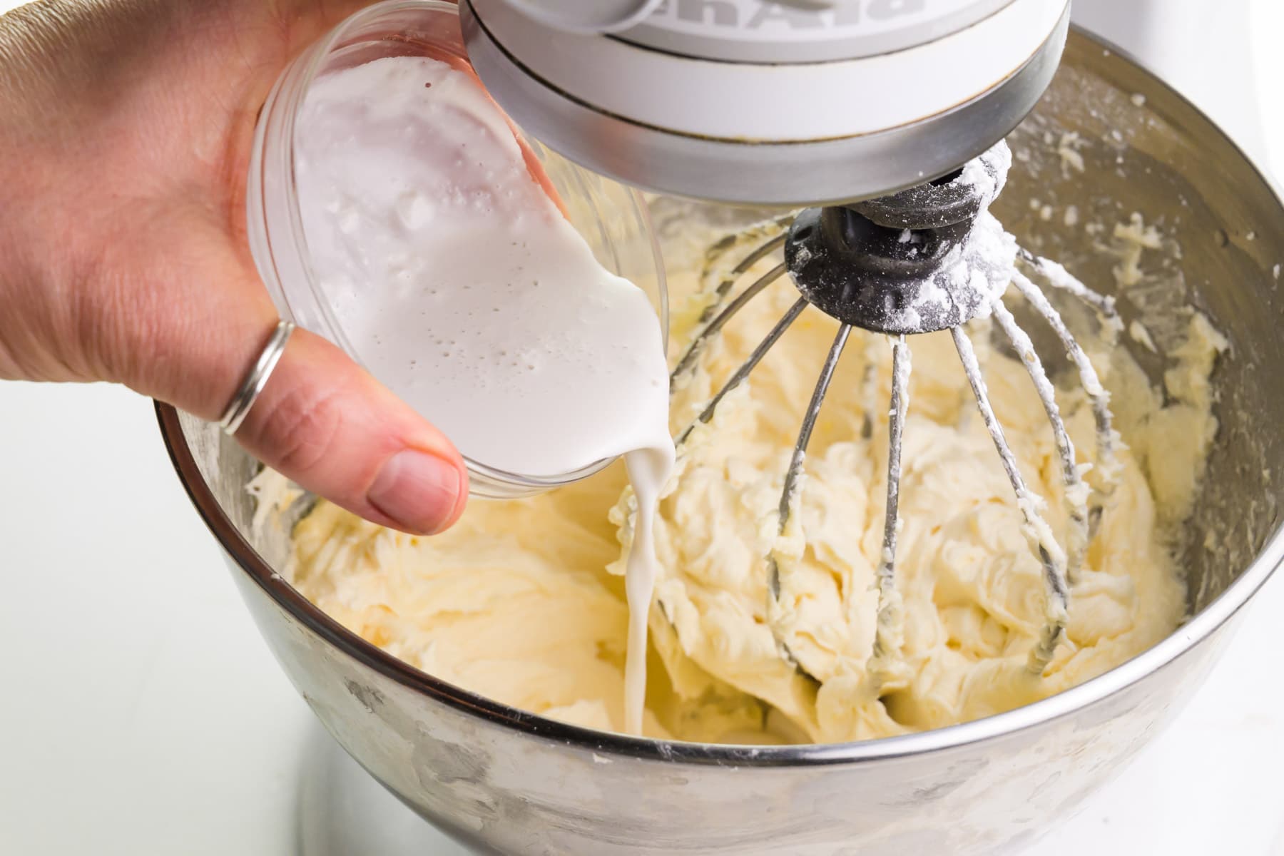 A hand holds a bowl of vegan creamer, pouring it into a stand mixer with vegan lemon frosting.