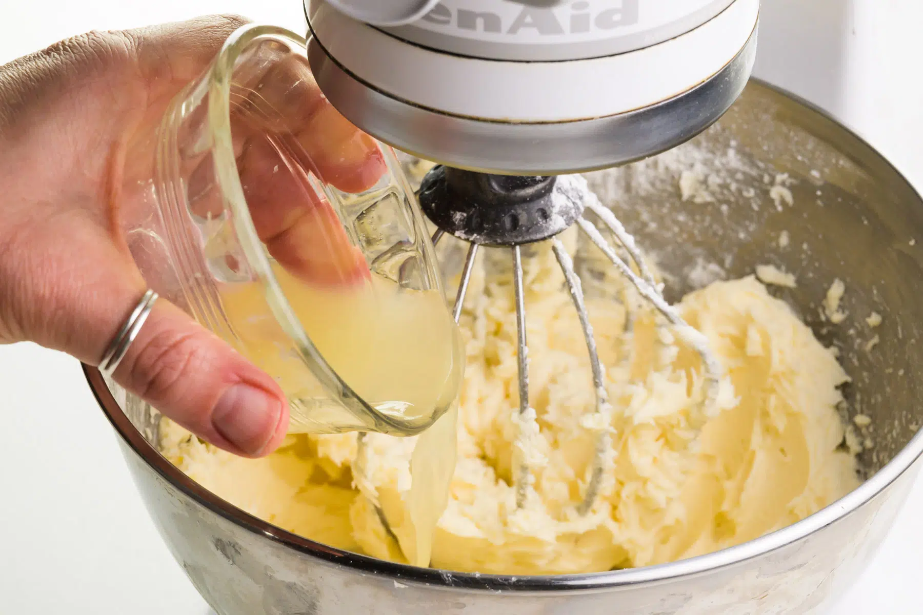 A hand holds a bowl of lemon juice, pouring it into the bowl of a stand mixer with whipped butter.