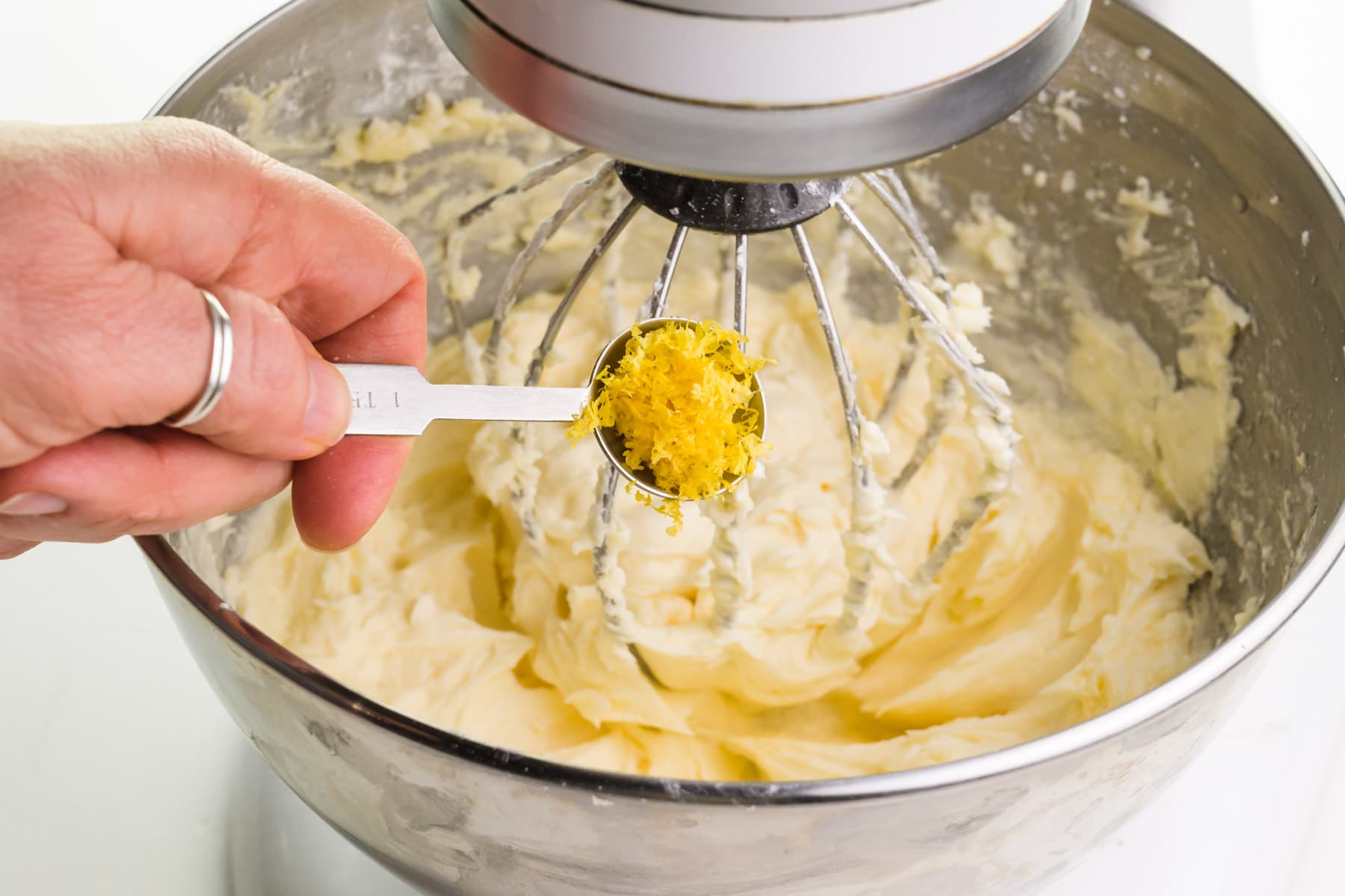 A hand holds a measuring spoon with lemon zest, pouring it into a stand mixer with whipped butter.