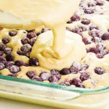 Batter is being poured over a lemon cake batter with blueberries on top.