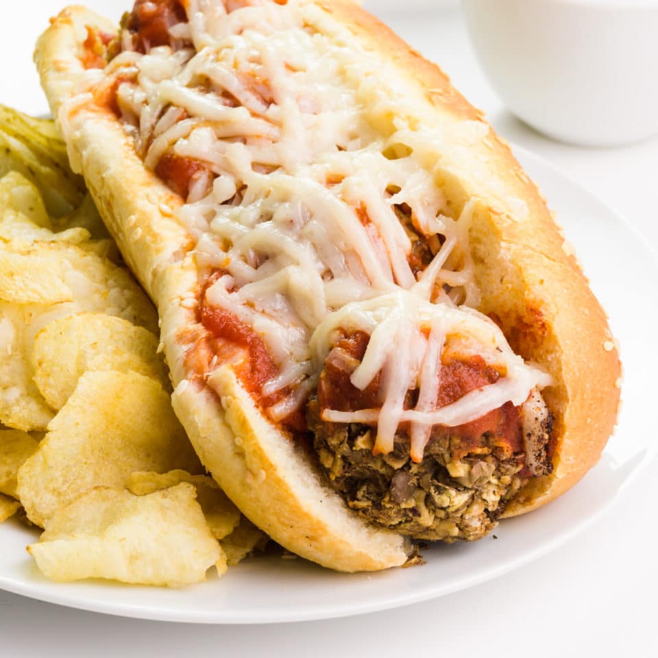 A vegan meatball sub sits on a plate next to potato chips.