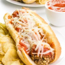 A vegan meatball sub sits on a plate next to potato chips. Behind it is a bowl of marinara and another sub on a plate.