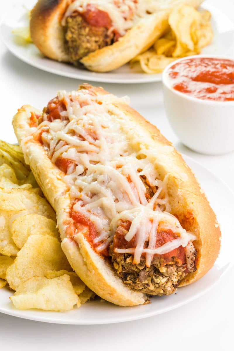 A vegan meatball sub sits on a plate next to potato chips. Behind it is a bowl of marinara and another sub on a plate.