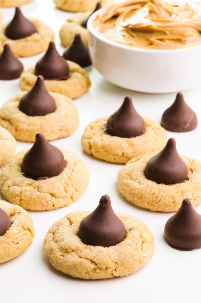 Several eggless peanut butter blossoms sit next to a bowl with peanut butter.