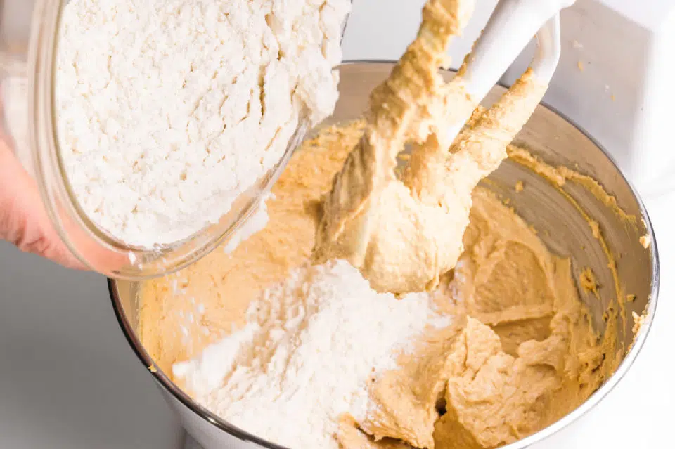 Flour is being poured into a mixing bowl with a whipped peanut butter mixture.