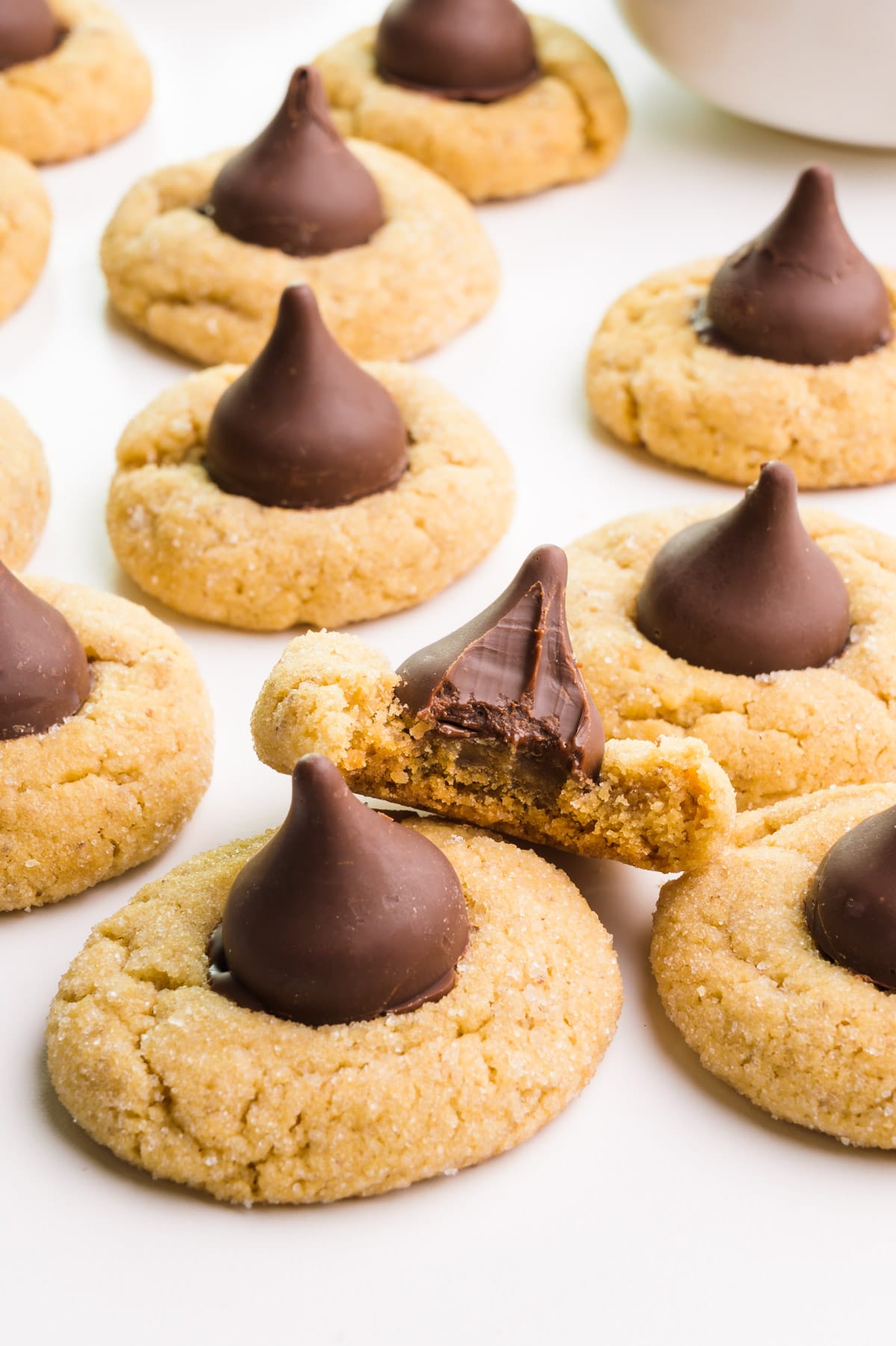 Several vegan peanut butter blossoms cookies sit on a white table. One has a bite taken out.