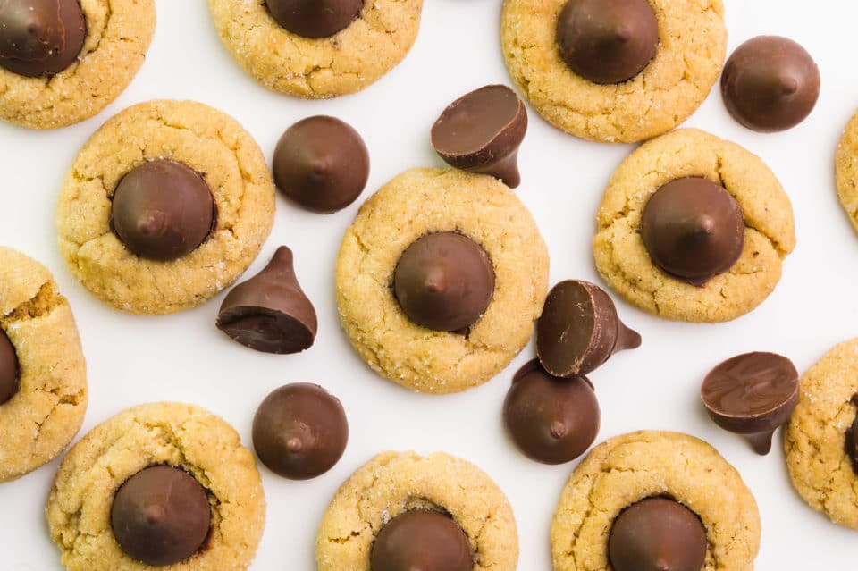 Below are several peanut butter cookies with chocolate kisses on top.  There are several vegan kiss candies around cookies.