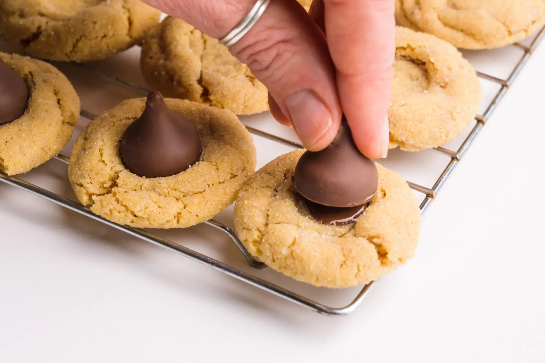 A hand holds a vegan chocolate kiss and is pressing it into the center of peanut butter cookies.