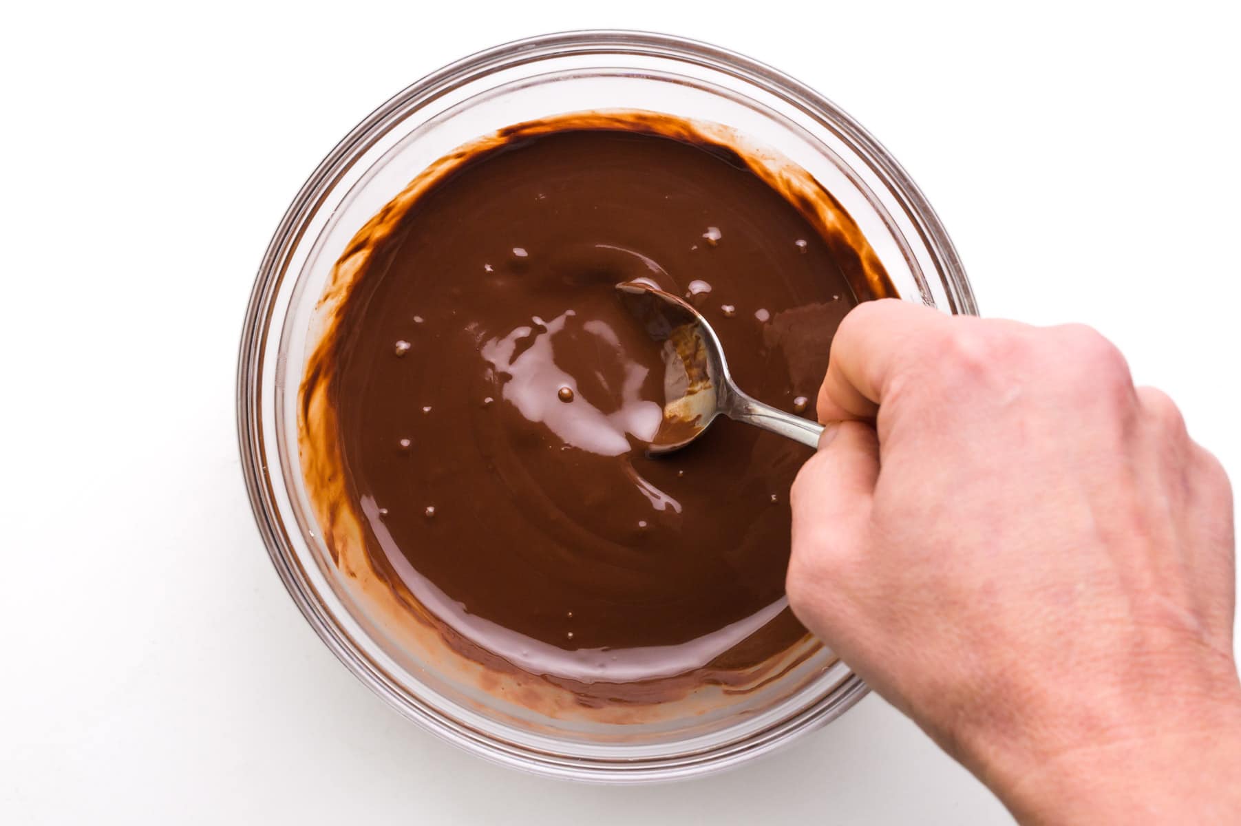A hand holds a spoon stirring melted chocolate in a bowl.
