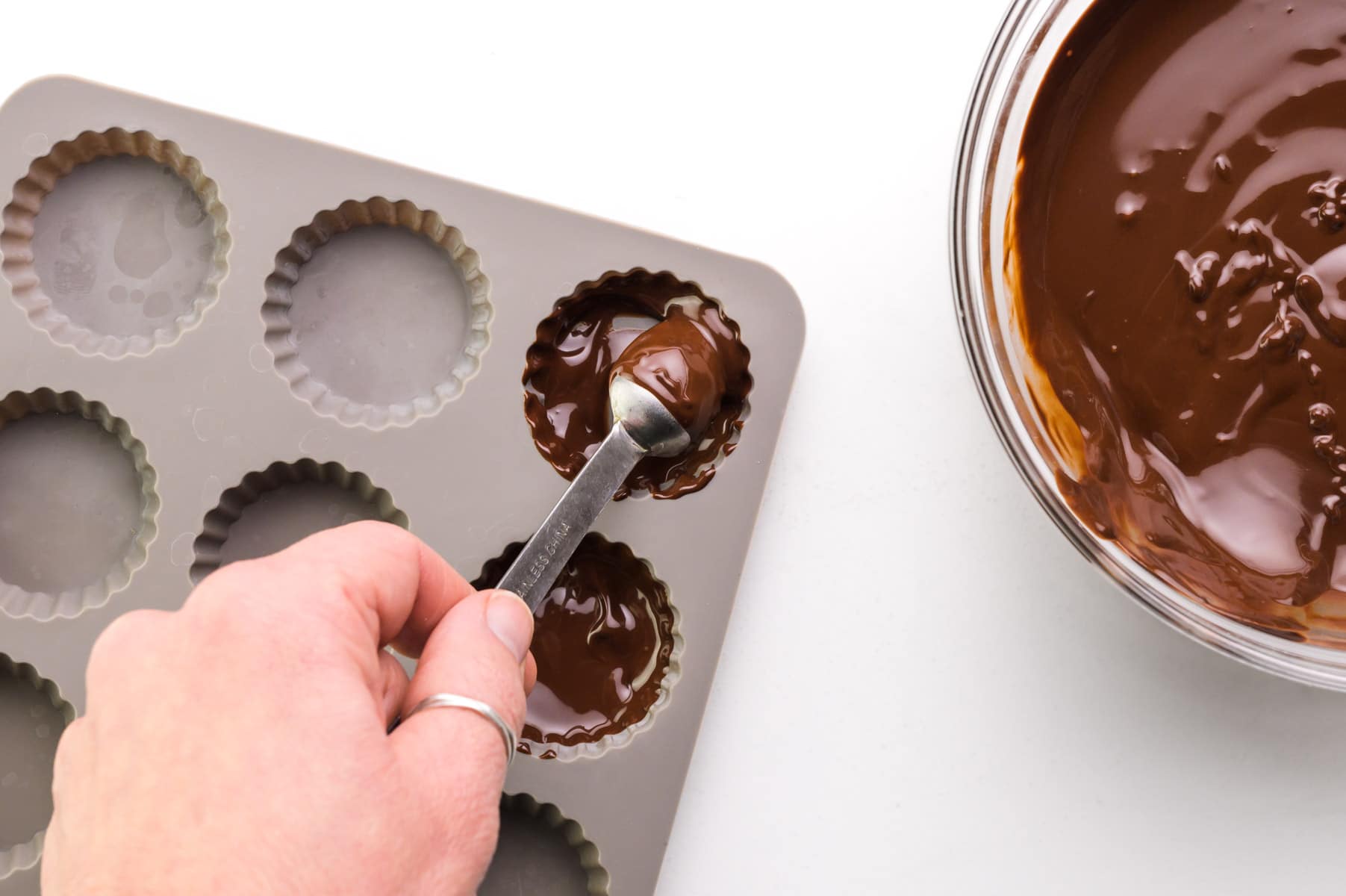 A hand holds a spoon spreading melted chocolate in a candy mold.