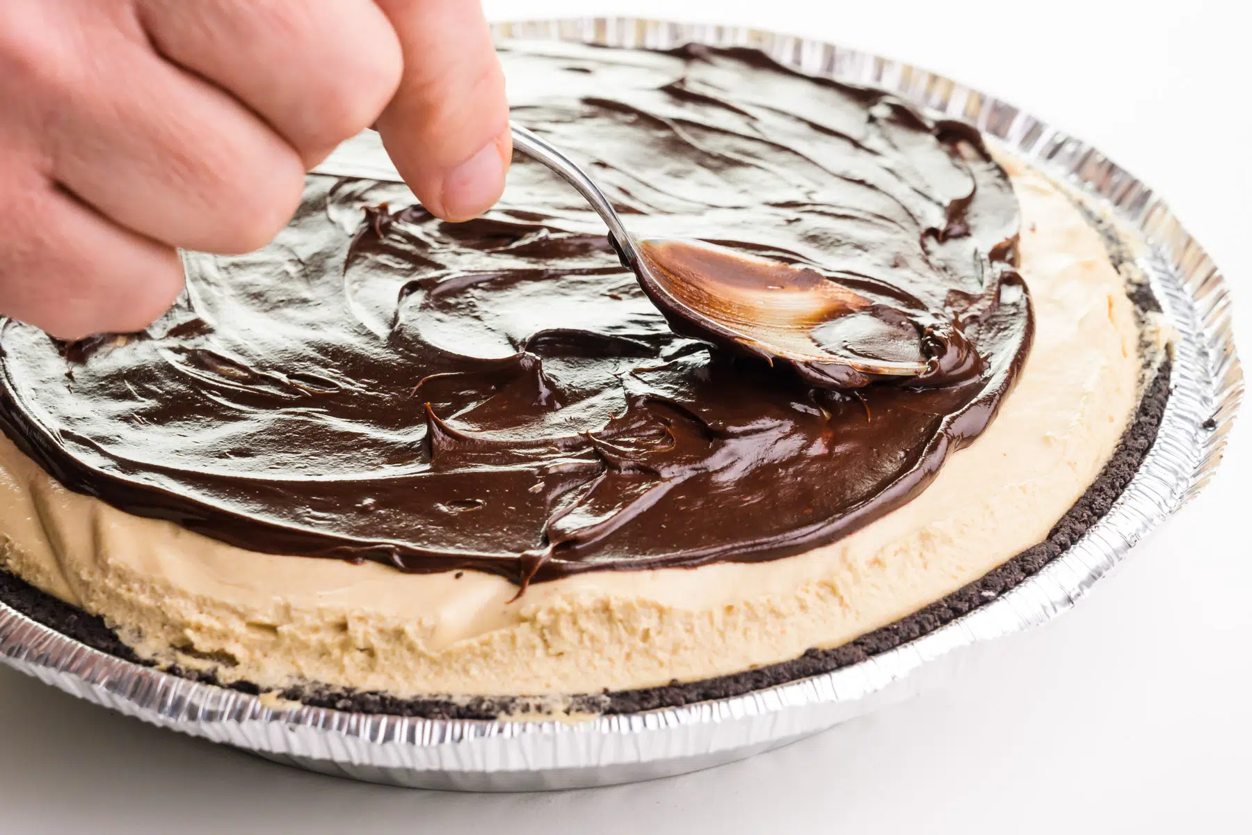 A hand holds a spoon, spreading out a chocolate topping on top of a peanut butter pie.
