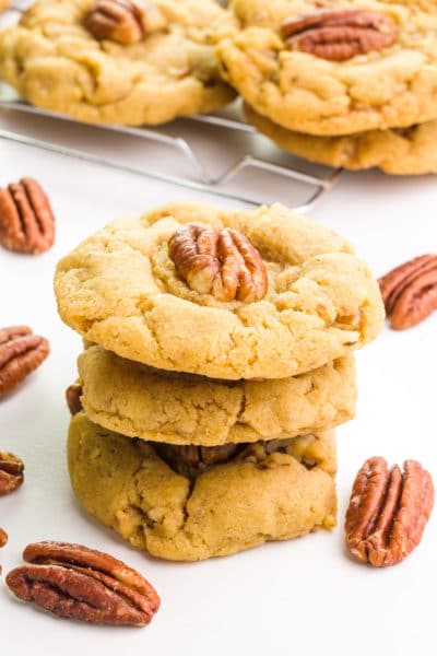 Vegan pecan cookies are stacked up with pecans around them and more cookies in the background.